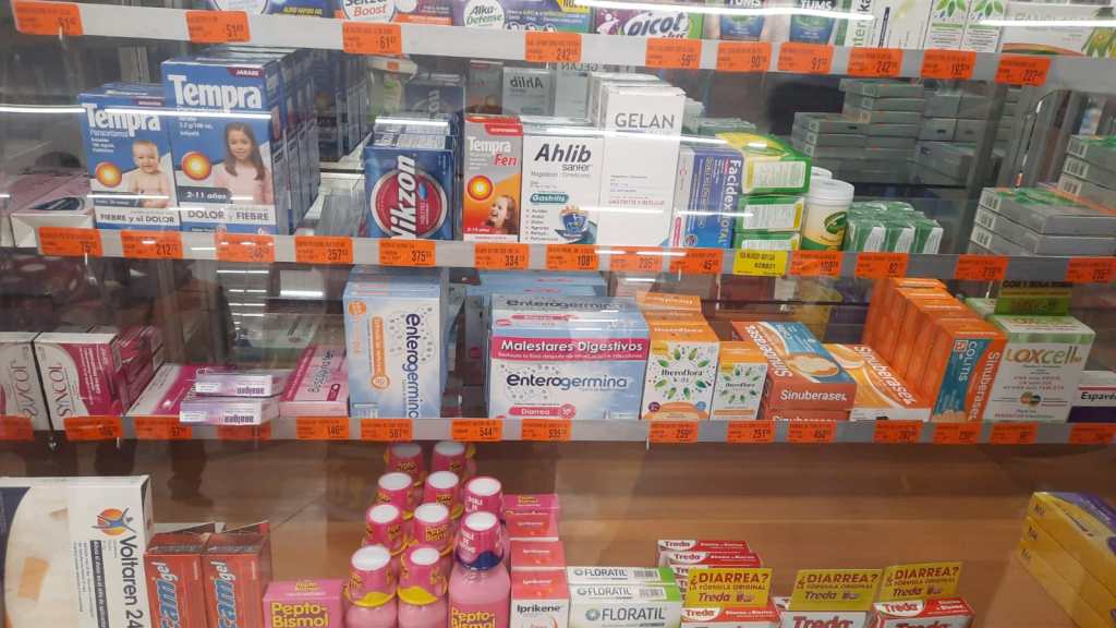 Estante de farmacia que muestra una variedad de medicamentos para el tratamiento de la gripe y otros síntomas, con precios en etiquetas naranjas.