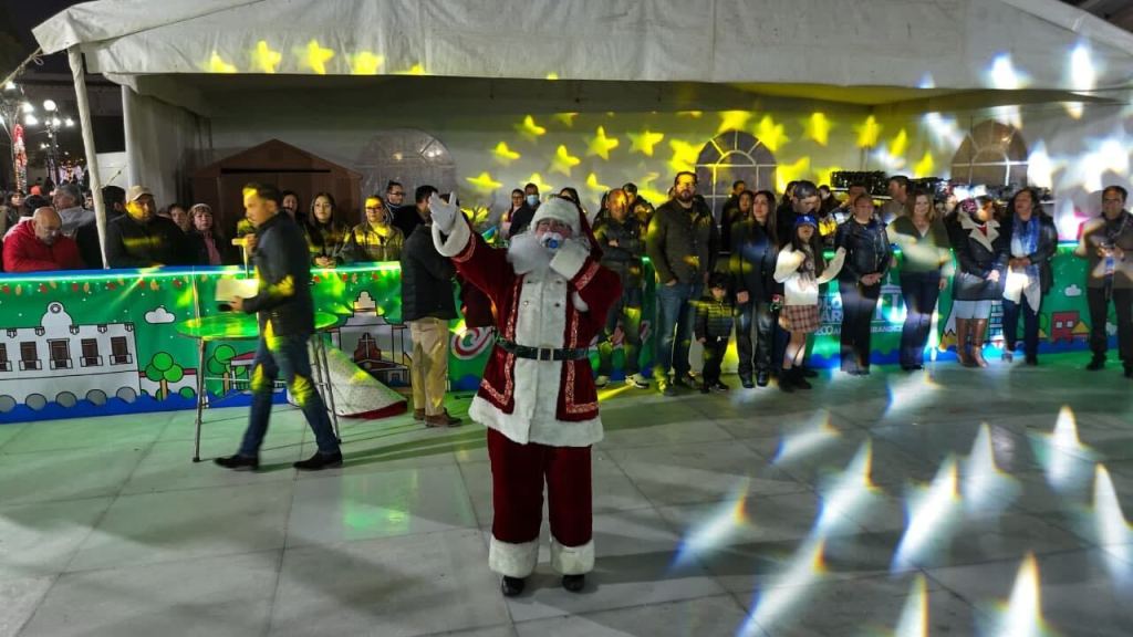 Un hombre vestido de Santa Claus saluda al público durante la inauguración de una ecopista de patinaje en Almoloya de Juárez. Al fondo, se ve a una multitud de personas reunidas, con luces decorativas y un ambiente festivo.