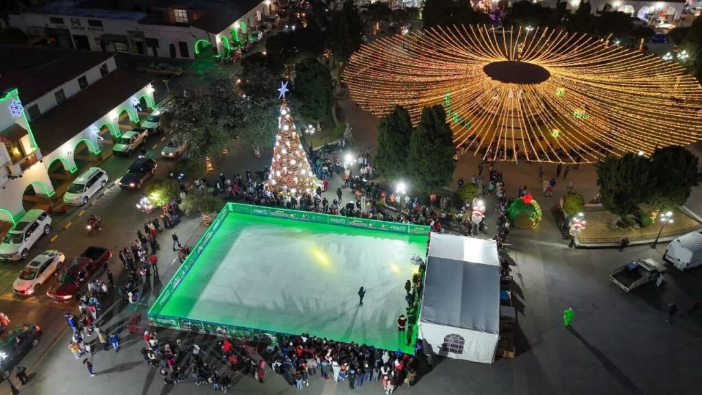 Vista aérea de una pista de patinaje ecopista en la explanada central de Almoloya de Juárez, rodeada de luces navideñas y un árbol de Navidad iluminado, con una multitud de personas esperando para patinar.