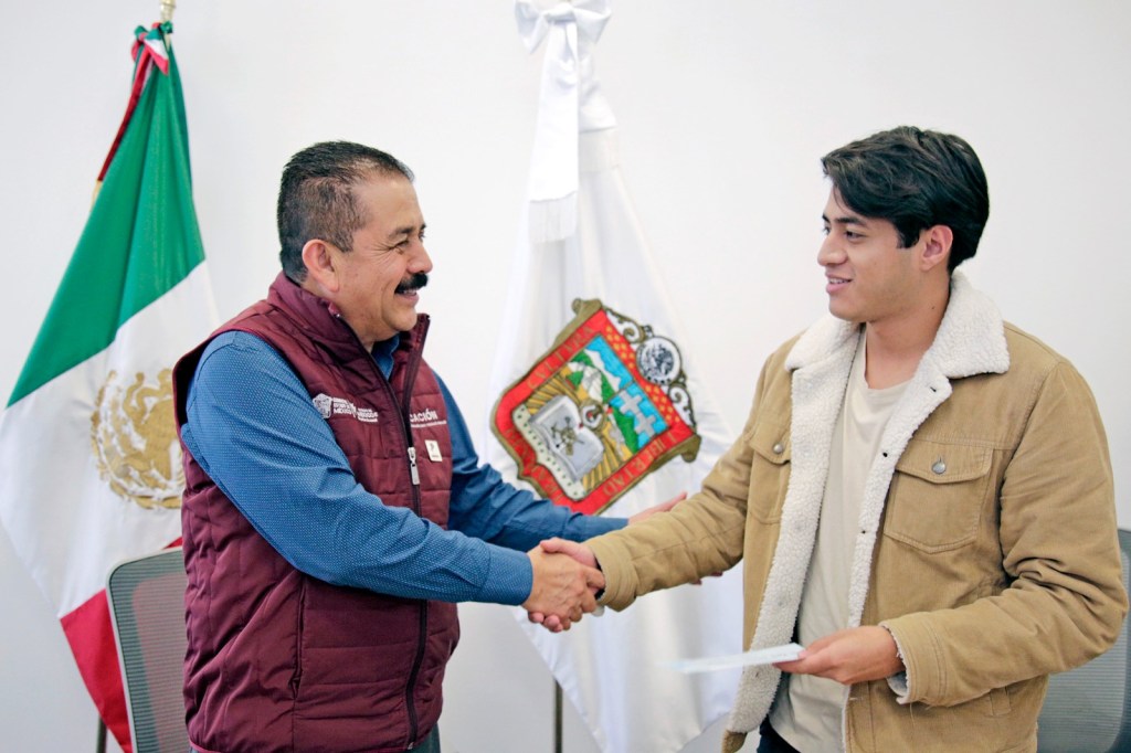 Un funcionario y un joven estrechan manos en un evento formal, con banderas de México y del Estado de México en el fondo.