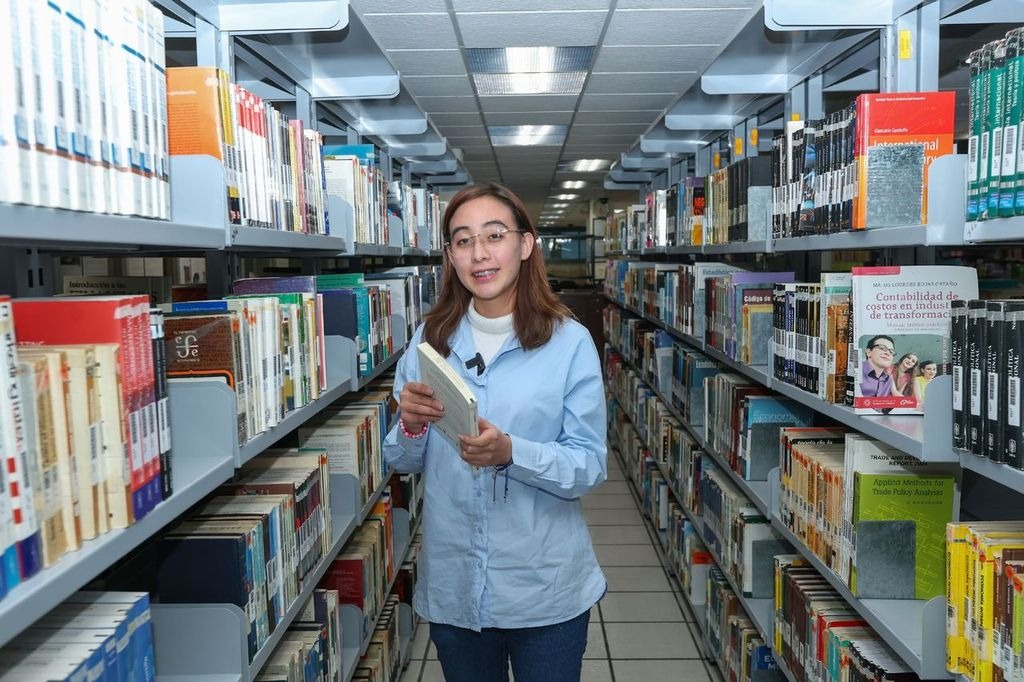 Una joven sostiene un libro en una biblioteca, rodeada de estanterías llenas de libros en diversos colores y tamaños.