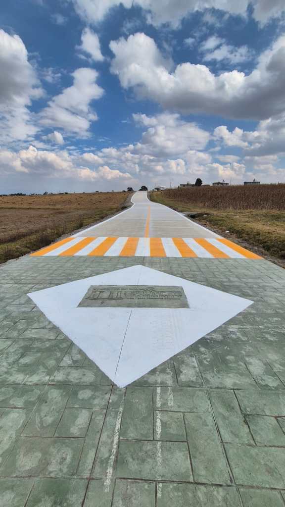 Caminos rehabilitados en Almoloya de Juárez, mostrando un camino de concreto hidráulico con un diseño en el pavimento, en un paisaje rural con nubes.