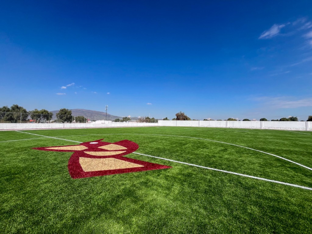 Campo de futbol con pasto sintético en la nueva Unidad Deportiva de Cuautlacingo, Estado de México, con un diseño en el centro y un cielo azul despejado.