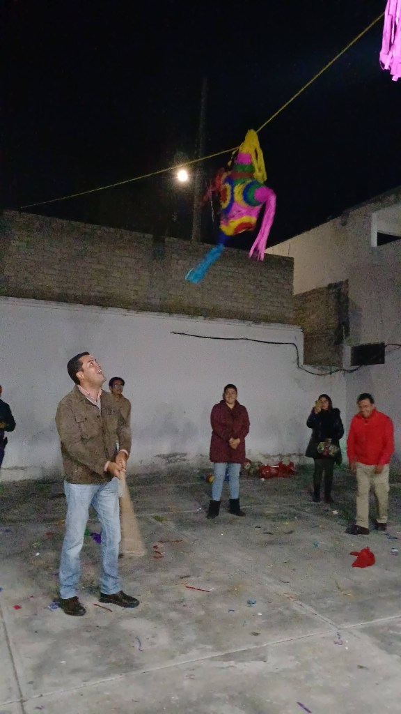 Un hombre está a punto de golpear una piñata colorida que cuelga sobre un fondo oscuro. A su alrededor, hay varias personas observando y celebrando en un ambiente festivo.
