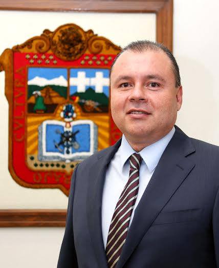 Retrato de un hombre de mediana edad con traje oscuro y corbata, sonriente, frente a un escudo enmarcado.