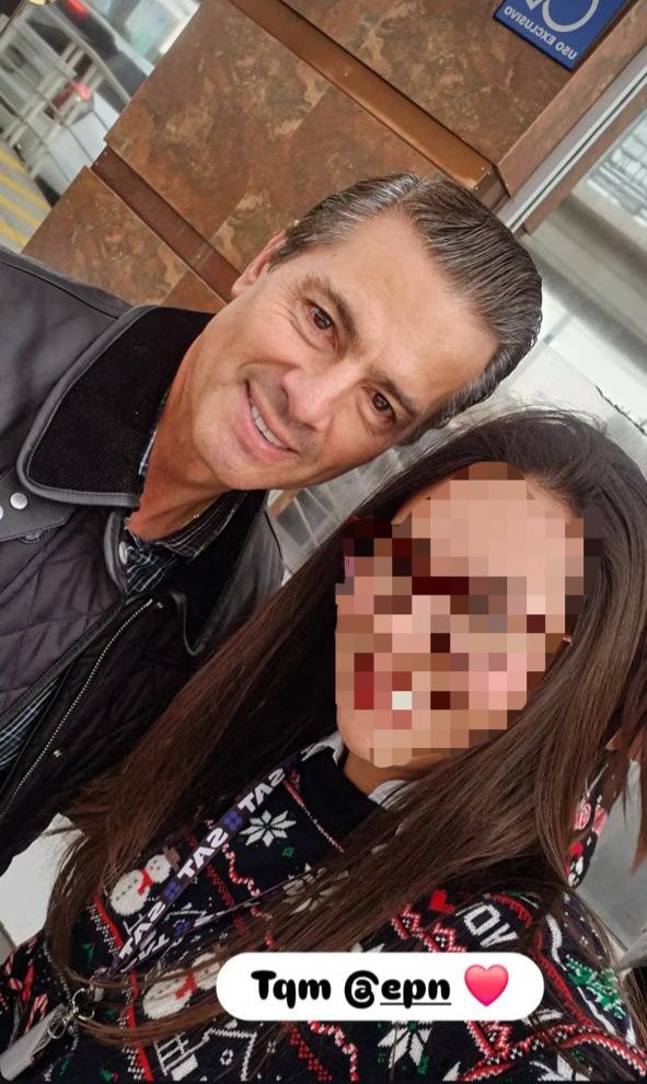 Enrique Peña Nieto posando para una selfie con una mujer en un entorno urbano, ambos sonriendo. La mujer lleva un suéter decorado con un diseño navideño.