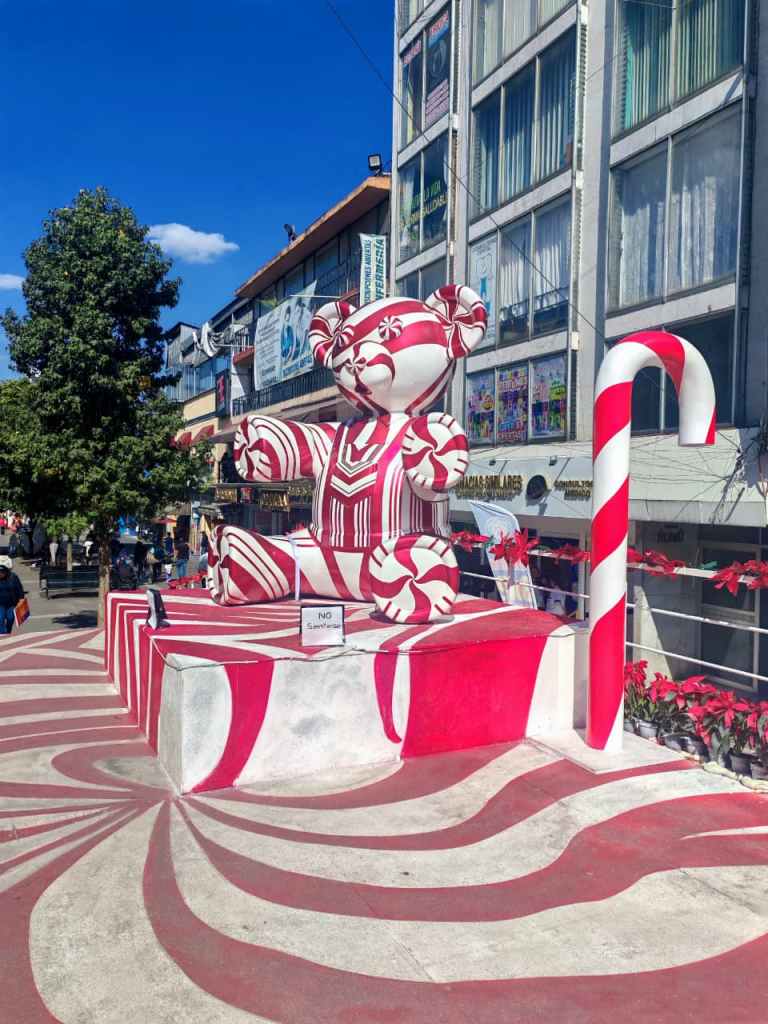 Escultura de un oso con temática navideña en tonos rojo y blanco, rodeado de decoraciones festivas y caramelos en un ambiente urbano.