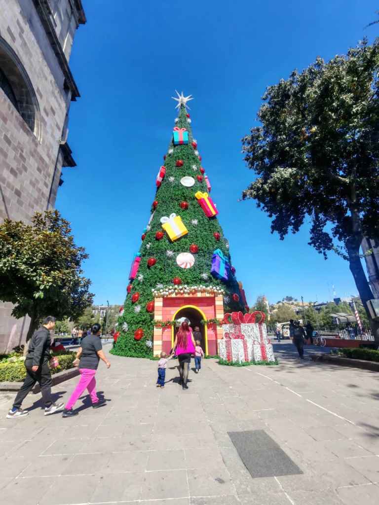 Árbol de Navidad decorado con regalos y adornos en un espacio público, con personas caminando cerca bajo un cielo azul.