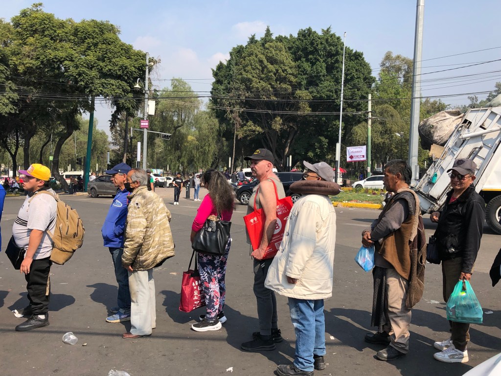 Un grupo de peregrinos en fila esperando llegar a la Basílica de Guadalupe en la Ciudad de México, con árboles y vehículos al fondo.