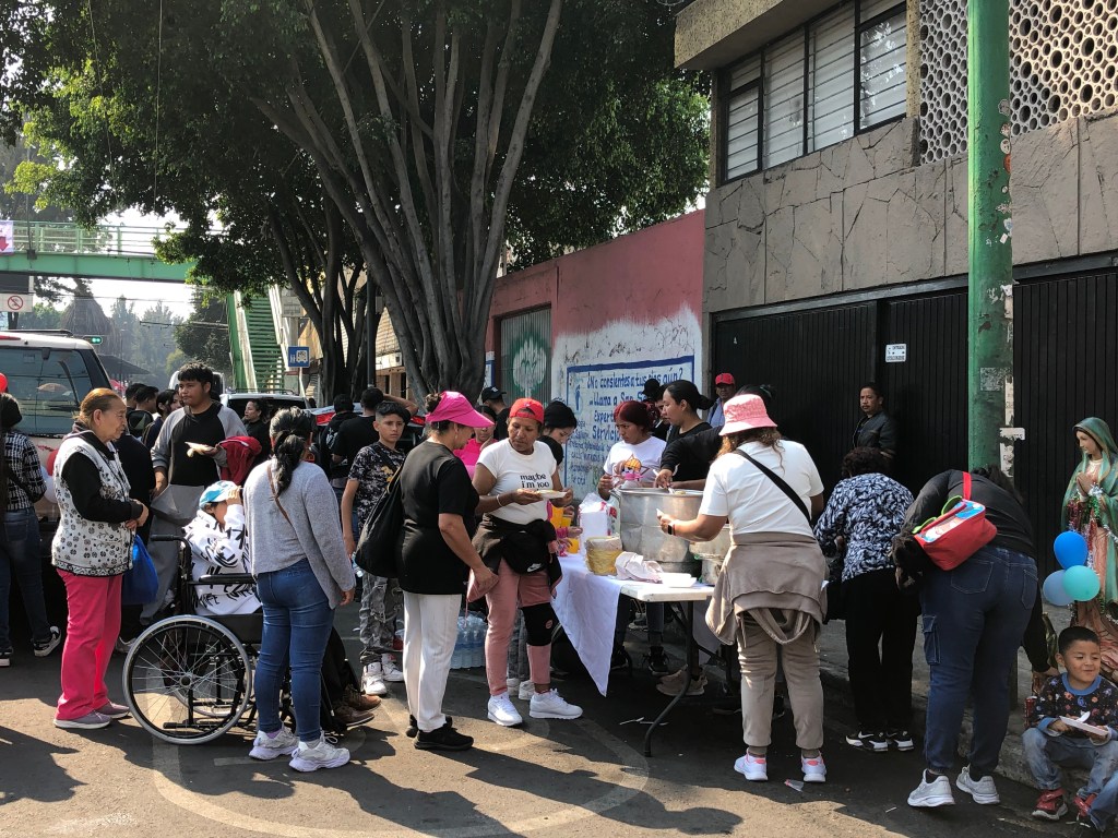 Grupo de personas reunidas en la calle, compartiendo alimentos y agua durante la celebración del 12 de diciembre en la Basílica de Guadalupe. Se pueden ver peregrinos, algunos con niños y otros en silla de ruedas, rodeados de árboles y carteles.