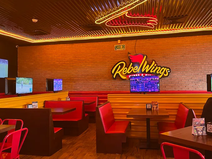 Interior de Rebel Wings, un restaurante con un ambiente casual, mesas y asientos rojos, pantallas para ver deportes y una decoración iluminada.