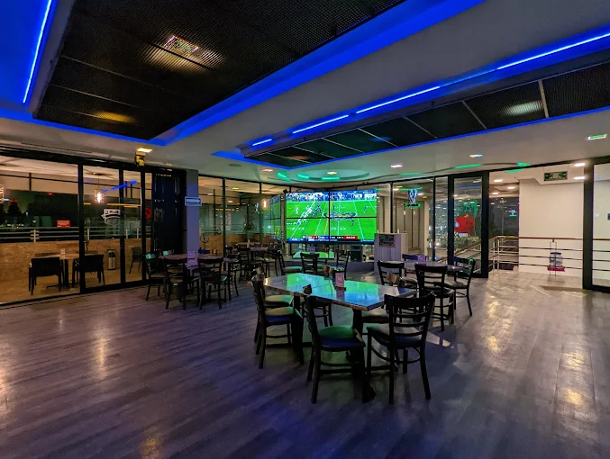 Interior de un restaurante con mesas y sillas, mostrando una gran pantalla donde se transmite un partido de fútbol.