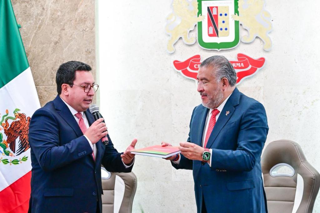 Horacio Duarte Olivares, Secretario General de Gobierno, entregando un documento a Miguel Ángel Ramírez Ponce, presidente municipal de Lerma, en un evento oficial con una bandera de México de fondo.