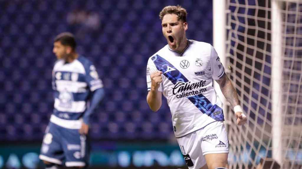 Un jugador de fútbol celebra tras anotar un gol, con una expresión de euforia mientras otros jugadores están en el fondo en medio de un partido.