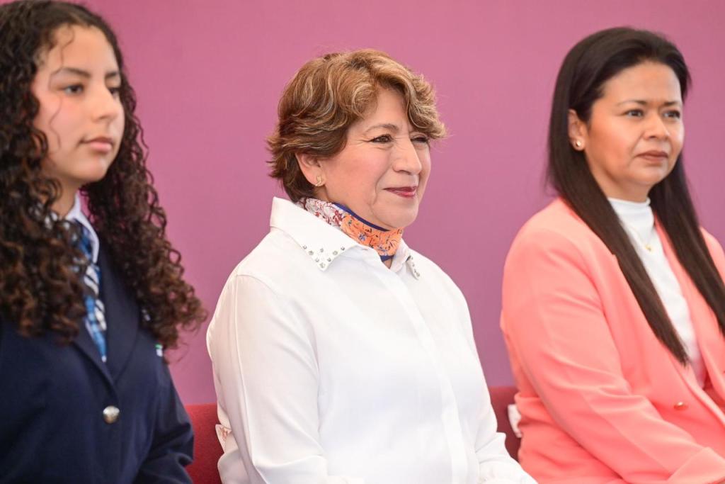 Tres personas sentadas en un evento, una de ellas posiblemente es la gobernadora, con un fondo morado.