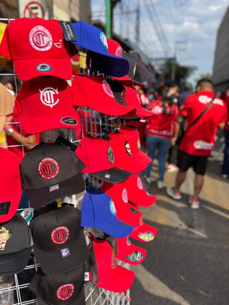Gorras rojas y negras del Club Deportivo Toluca exhibidas en un puesto, con varios fanáticos vestidos de rojo en el fondo.