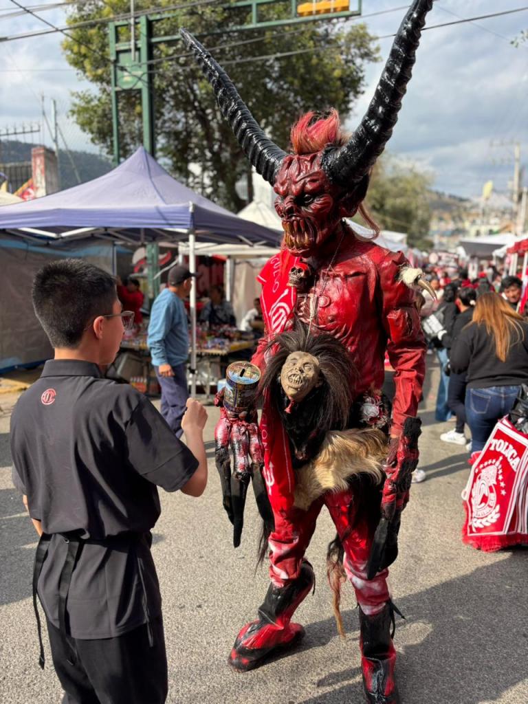 Un niño interactúa con un hombre disfrazado de diablo, con cuernos y un traje rojo, en un ambiente festivo previo a un partido de fútbol.