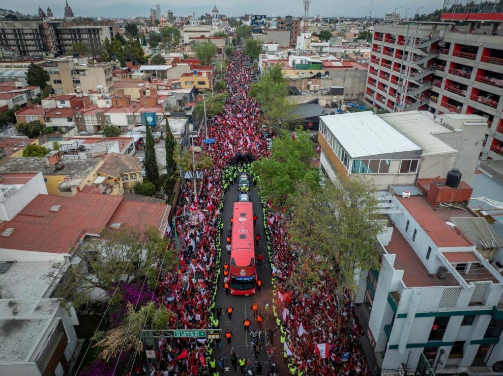 Vista aérea de una multitud de aficionados vestidos de rojo en las calles, acompañando el autobús de los Diablos Rojos del Toluca hacia el Estadio Nemesio Diez.