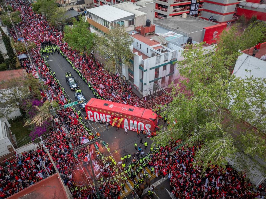 Aerial view of a large crowd of fans dressed in red, lining the streets to welcome the Toluca FC team bus, with security personnel present and festive decorations.