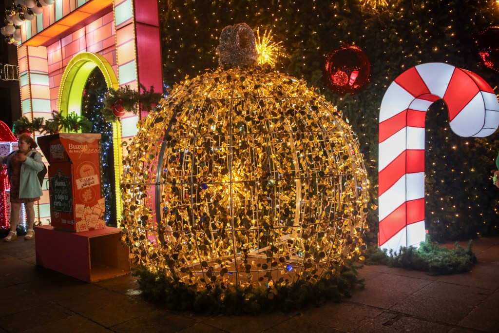 Esfera navideña iluminada decorada con luces doradas, con un fondo de elementos festivos como un bastón de caramelo y una figura decorativa, creando un ambiente navideño en el centro de Toluca.