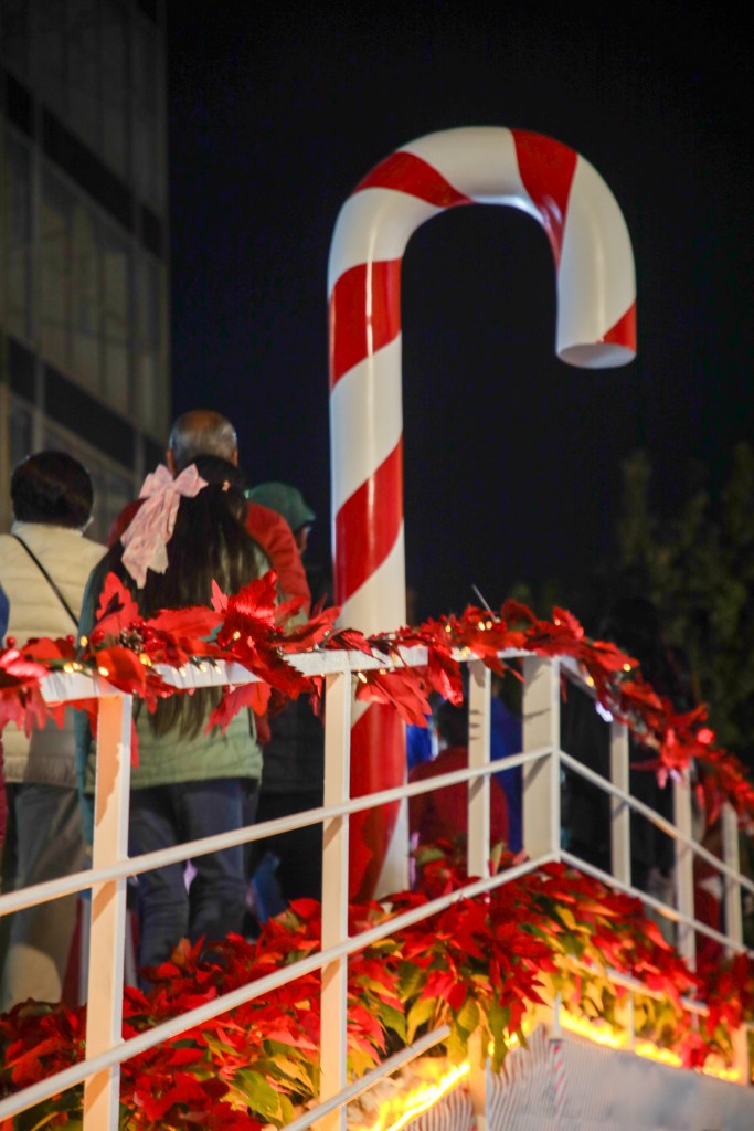 Un gran bastón de caramelo decorativo, rodeado de flores rojas y un grupo de personas disfrutando del ambiente navideño.