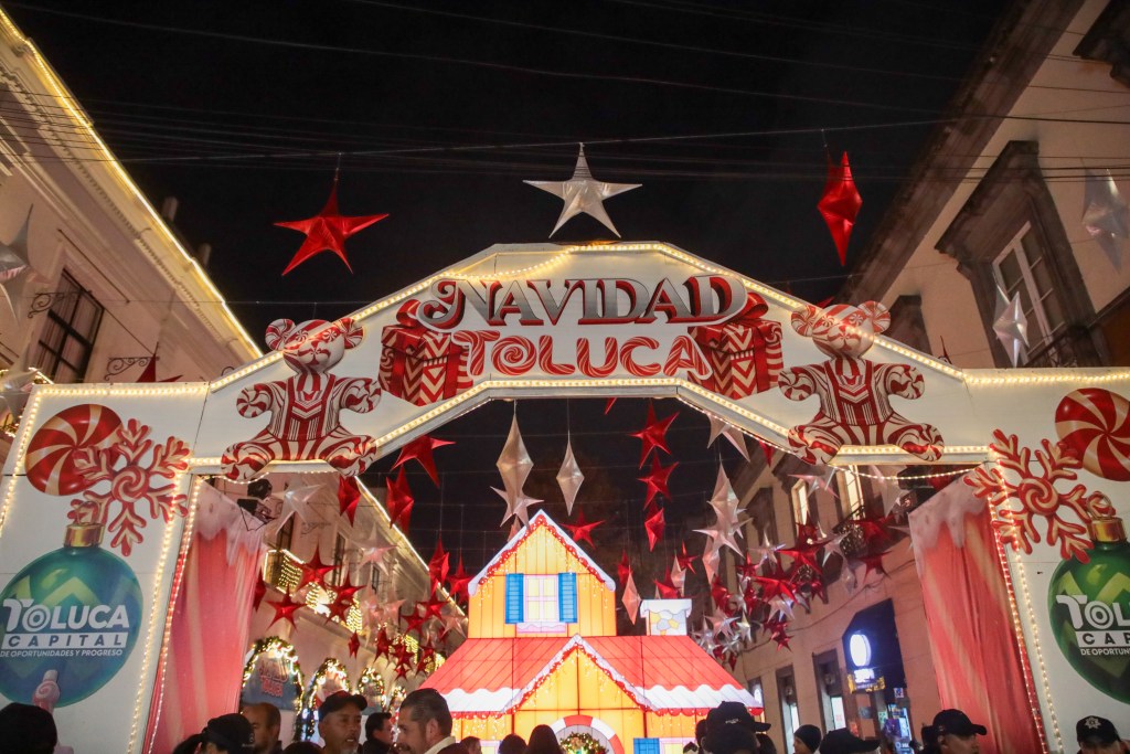 Arco decorativo con la frase 'Navidad Toluca' adornado con estrellas rojas y elementos navideños, junto a una casita navideña.
