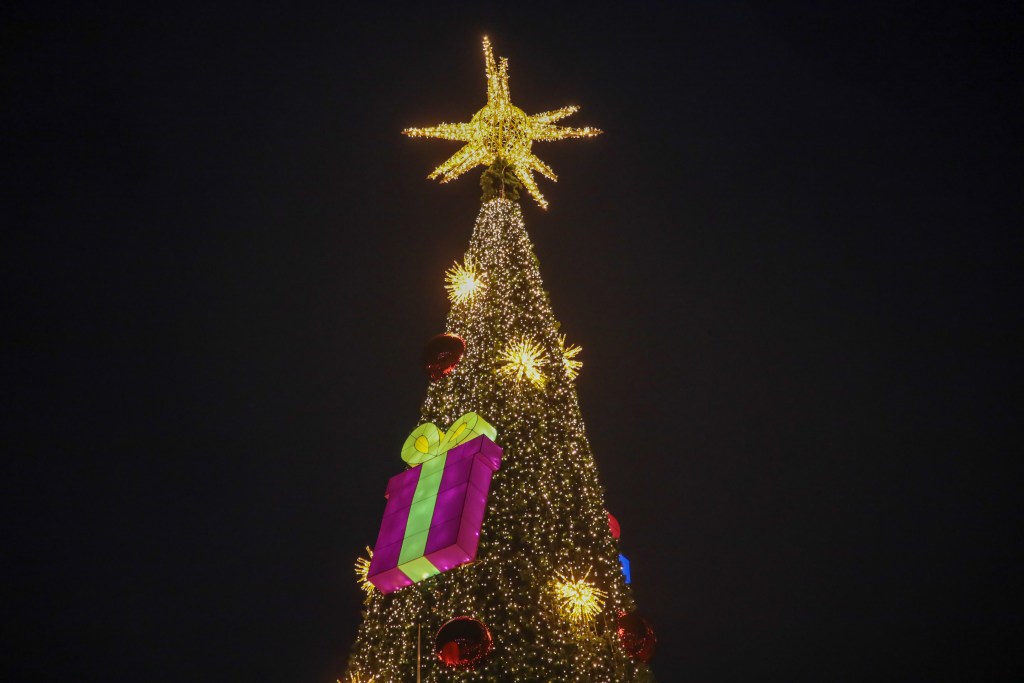 Gran árbol navideño iluminado con estrellas y adornos coloridos, incluyendo un regalo gigante en la base.