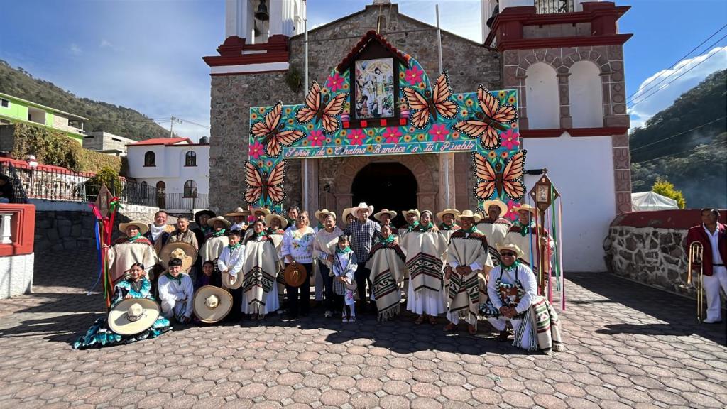Grupo de danzantes con trajes tradicionales frente a una iglesia decorada con mariposas y un arco colorido en la comunidad de Santiago Analco.
