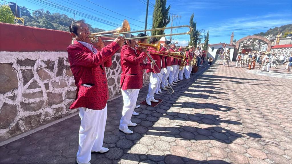 Trombonistas en un evento cultural, vestidos con chaquetas rojas y pantalones blancos, tocando en una plaza al aire libre.