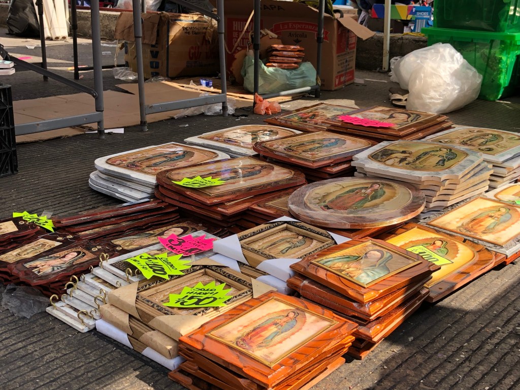Puestos ambulantes con imágenes religiosas de la Virgen de Guadalupe exhibidas en el suelo, con precios marcados en etiquetas brillantes.