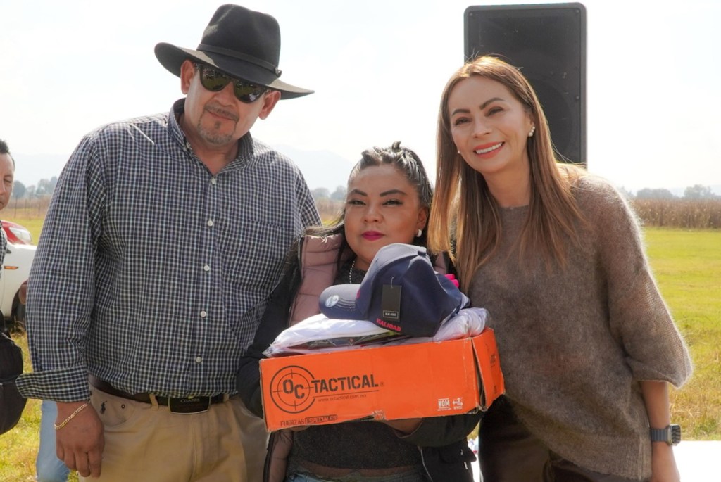 Tres personas posando en un evento de entrega de uniformes, una de ellas sostiene un paquete con una gorra mientras sonríen.