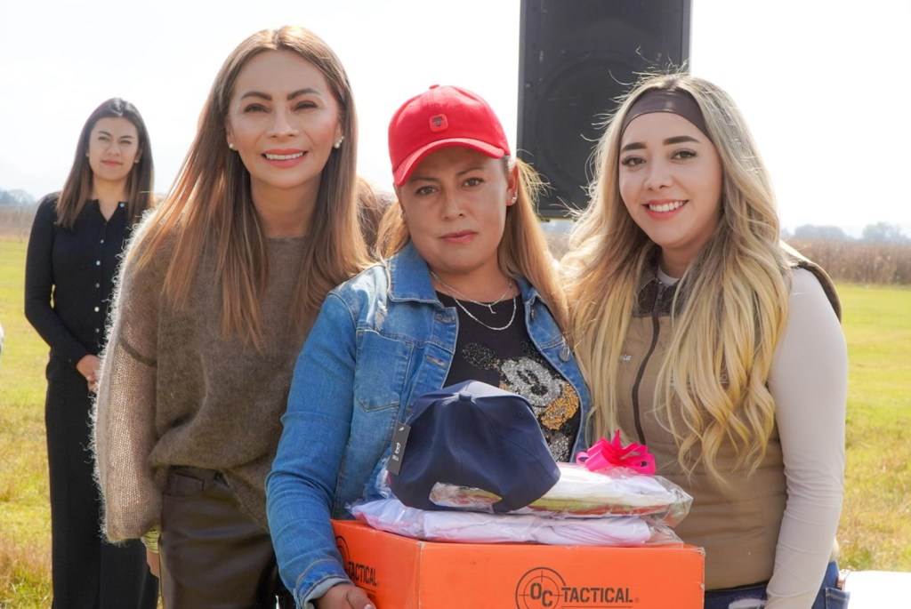 Tres mujeres sonrientes posan para la foto, una de ellas sostiene una caja de uniformes. Se encuentran al aire libre, con un campo en el fondo y un altavoz visible.