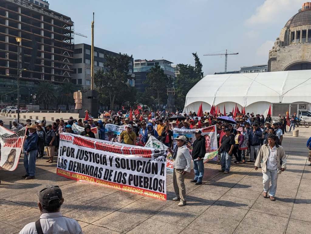 Manifestación en la que participan diversas personas sosteniendo pancartas que exigen justicia y soluciones a las demandas sociales. Se observa un ambiente de protesta pacífica con banderas y carteles en un espacio público.
