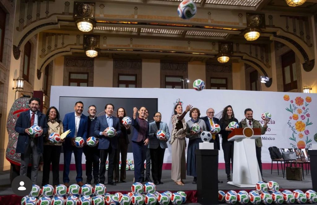 Un grupo de personas sostiene balones de fútbol durante un evento en un recinto formal en México, con decoraciones y un fondo que resalta la temática del Mundial 2026.jóvenes construyendo el futuro mundial 2026