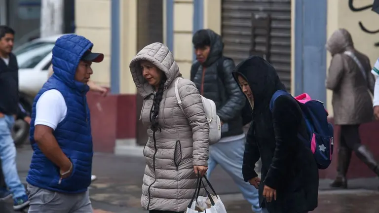 Grupo de personas caminando por la calle en un día frío, algunos con abrigos y capuchas para protegerse del clima. Se observan vehículos aparcados y edificios de fondo.