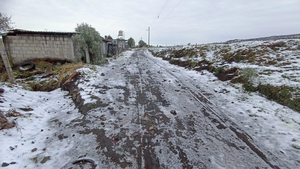 Un camino rural cubierto de hielo y nieve en una zona montañosa del Estado de México, con vegetación y edificaciones al costado.