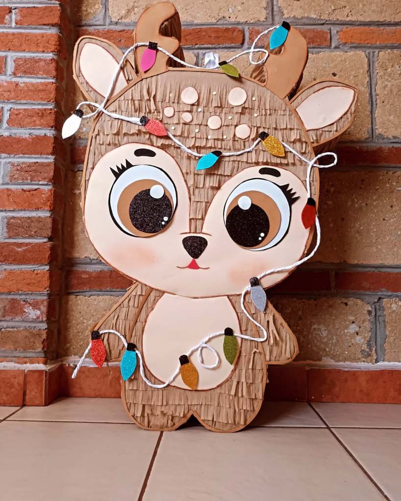 Piñata con forma de reno decorada con luces navideñas, apoyada en una pared de ladrillos.