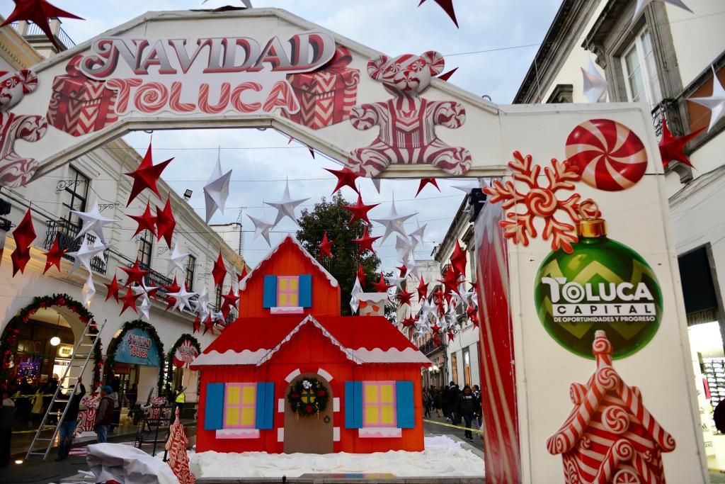 Arco decorativo de 'Navidad Toluca' en un evento festivo, con una casa navideña roja y decoraciones como estrellas y dulces en el decorado.