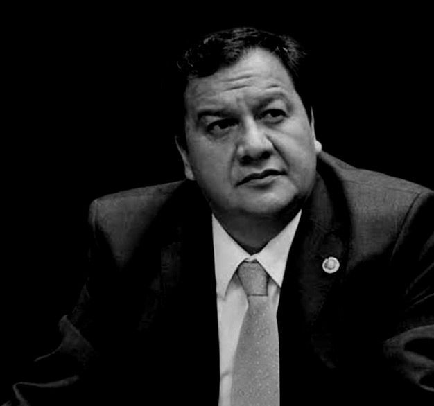 Retrato en blanco y negro de una persona con un traje oscuro, camisa clara y corbata, con una expresión seria y un fondo negro.
