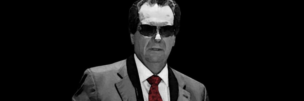 Hombre con traje gris, corbata roja y gafas oscuras, posando frente a un fondo negro.