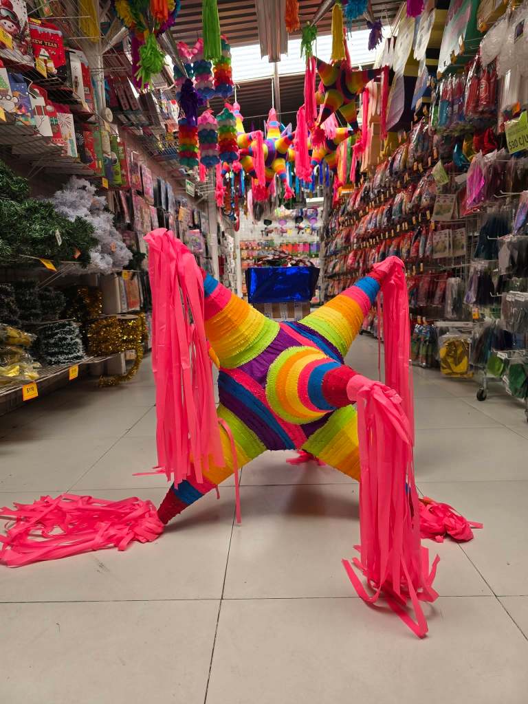 Una piñata colorida en forma de estrella con cintas rosas en un pasillo de una tienda de fiestas, rodeada de diversas decoraciones festivas.