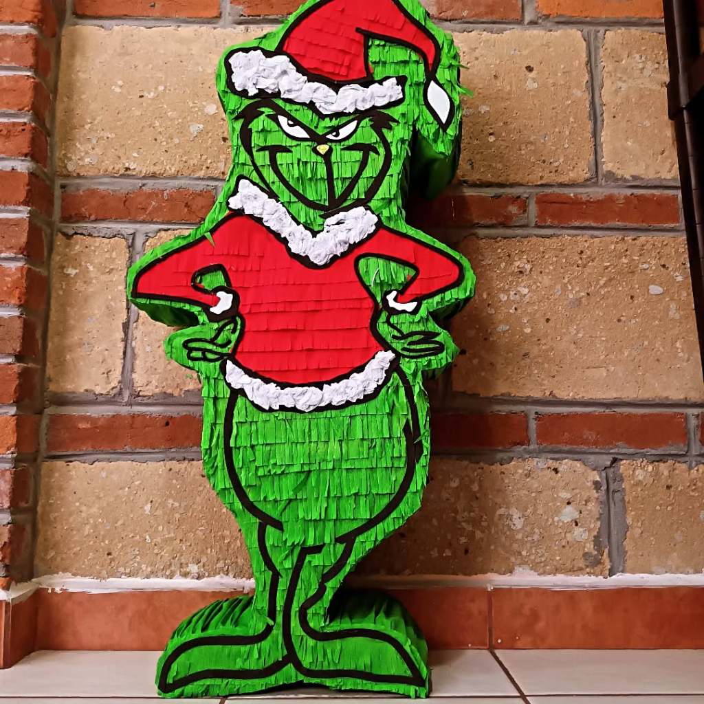 Piñata de Grinch en un fondo de pared de ladrillo, con detalles en rojo y blanco, lista para las celebraciones navideñas.