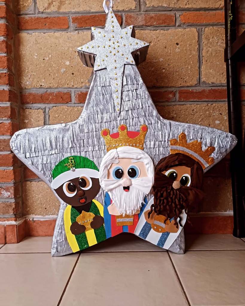 Piñata en forma de estrella decorada con figuras de personajes festivos, incluyendo un rey con corona, un rey con barba y un rey en vestimenta tradicional, sobre un fondo de ladrillos y piso de cerámica.