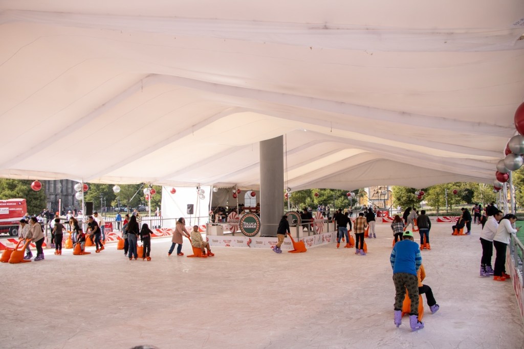 Vista de la pista de hielo en Toluca, con personas patinando y utilizando apoyos en un ambiente decorado para la temporada navideña.