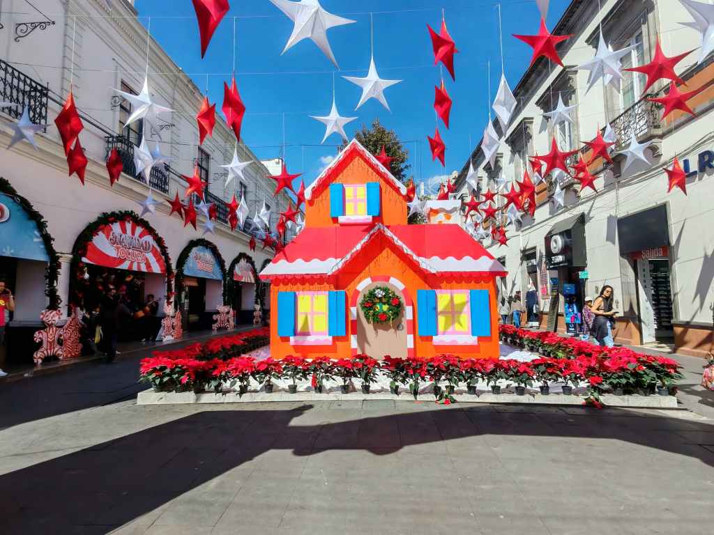 Casita de Jengibre decorada para Navidad, rodeada de flores de pascua y estrellas colgantes en la Plaza de los Mártires de Toluca.