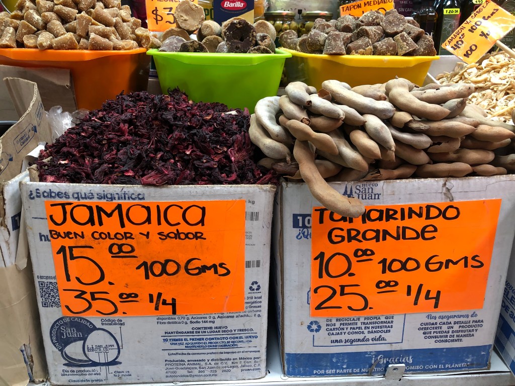 Caja de jamaica y tamarindo en un mercado, con precios visibles: jamaica a $15 por 100 g y $35 por un cuarto, tamarindo a $10 por 100 g y $25 por un cuarto.