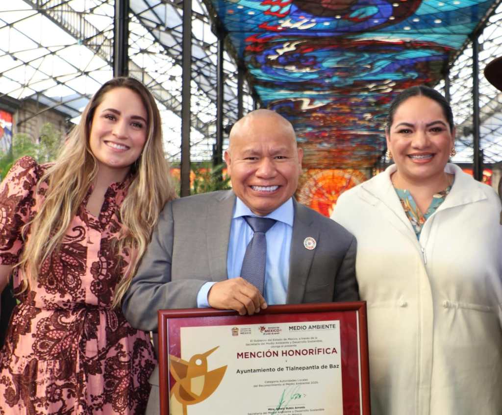 Tres personas posan sonrientes, sosteniendo un reconocimiento en un entorno con un techo decorado, que destaca por sus colores vibrantes.