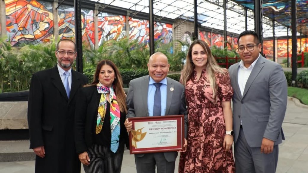 Grupo de personas posando en un espacio interior con plantas y arte mural, sosteniendo un reconocimiento honorífico.