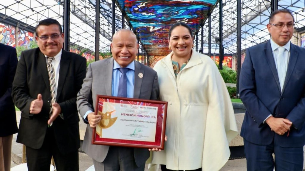 Grupo de personas sonrientes posando en un espacio al aire libre con una estructura de vidrio y coloridos vitrales. Una persona sostiene un diploma que dice 'Mención Honorífica' en español.