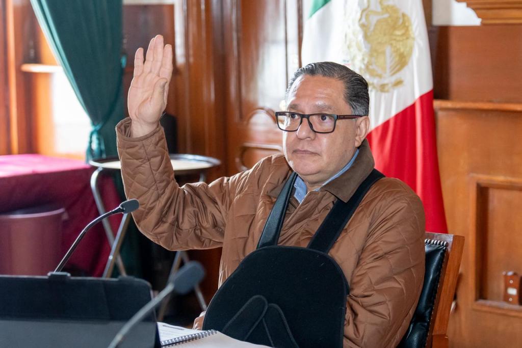 Un hombre con gafas y una chaqueta marrón levanta la mano durante una sesión del Ayuntamiento, con un fondo que incluye la bandera de México y cortinas verdes.